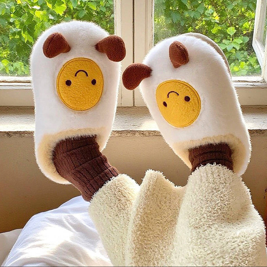 Pair of plush egg-shaped slippers with cartoon faces in front of a window with greenery.